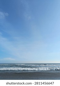 Continuous Ocean Waves Under an Open Sky. A clean vertical landscape shot displays multiple rows of frothy white waves rolling over the dark ocean surface beneath a vast, serene blue sky with faint, streaky clouds.