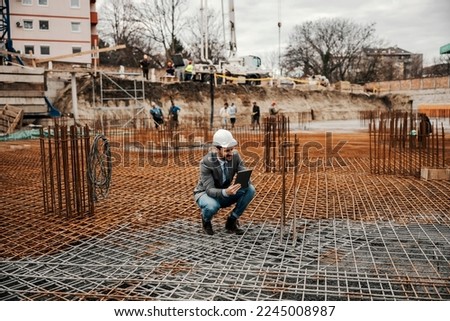 A contractor is crouching on building foundation at site and looking at the tablet.