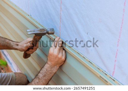 Contractor is using hammer to secure facade vinyl siding plank on exterior wall of house under renovation.
