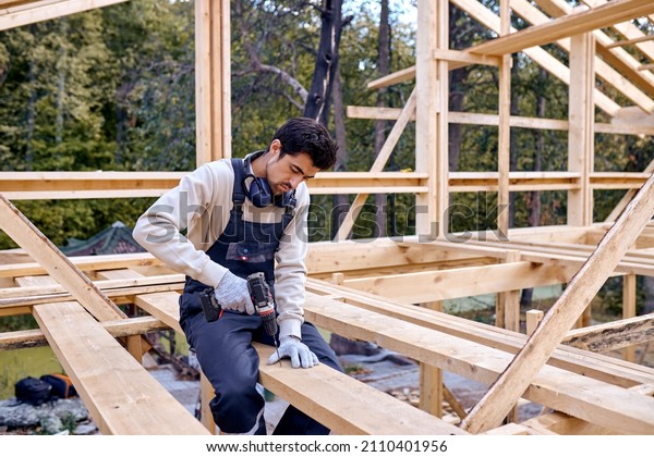 contractor in work clothes sit on beam install new\
roofing tools, using electric drill. Home renovation, sustainable\
living, building cottage house. Building Technologies. Industrial\
Theme.