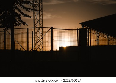 Contrast silhouette of an industrial building and lattice fence against the sunset yellow brown sky. Warehouse, factory building roof. Commercial property exterior. Wire fence in industrial zone. - Powered by Shutterstock