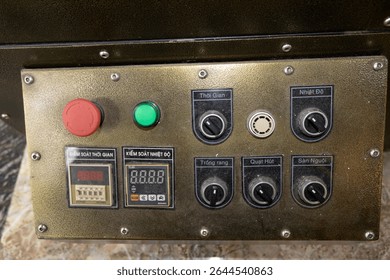 A control panel with a red emergency stop button, green start button, digital display, and multiple black knobs and switches on a metallic surface.