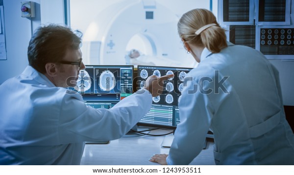 In\
Control Room Doctor and Radiologist Discuss Diagnosis while\
Watching Procedure and Monitors Showing Brain Scans Results, In the\
Background Patient Undergoes MRI or CT Scan\
Procedure.