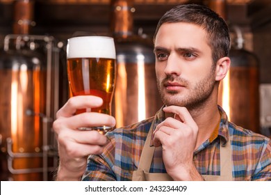 Controlling beer quality. Thoughtful young male brewer in apron holding glass with beer and looking at it while standing in front of metal containers - Powered by Shutterstock