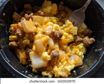 Convenience Meal Breakfast Scramble Featuring Sausage, Potatoes, Scrambled Eggs, And Cheddar Cheese In A Black Microwavable Bowl With A Fork 