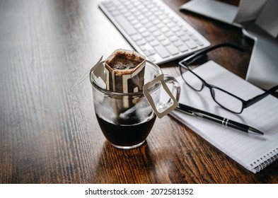 Convenient Paper Drip Coffee Bag In Glass Cup On Dark Office Table. Making Freshly Brewed Coffee At Work