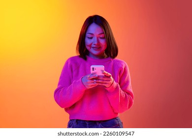 Conversation  Young girl in pink sweater typing text message mobile phone over gradient orange background in neon light  Concept emotions  facial expression  youth  inspiration  sales  ad