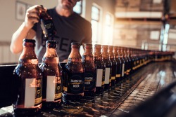 Przenośnik Z Butelkami Piwa W Fabryce Browaru. Młody Człowiek Nadzorujący Proces Butelkowania Piwa I Sprawdzający Jakość W Zakładzie Produkcyjnym.