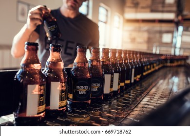 Conveyor with beer bottles moving in brewery factory. Young man supervising the beer bottling process and checking the quality at the manufacturing plant. - Powered by Shutterstock