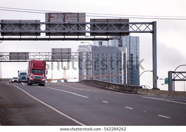 Convoy of\
big rigs American semi trucks with various types of trailers moving\
along overpass wide highway which passing by the big city, the\
designated apartment multistory\
buildings