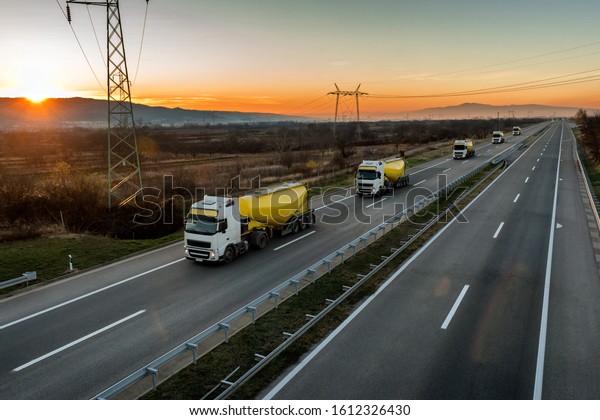 Convoy or\
Caravan of white and yellow Tank trucks on a Highway traffic\
through the rural landscape at beautiful\
sunset