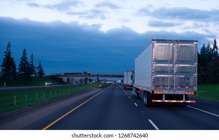 The Convoy Of Semi Trucks With Reefer Trailers On Flat Like An Arrow Evening Road With Lights On And Reflection Of Light On A Shiny Trailer Stainless Steel Doors. Trucks Driving On A Divided Highway