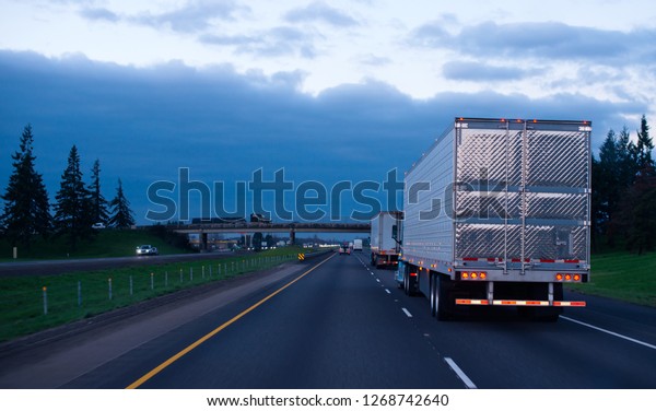 The convoy of semi trucks with reefer trailers on\
flat like an arrow evening road with lights on and reflection of\
light on a shiny trailer stainless steel doors. Trucks driving on a\
divided highway