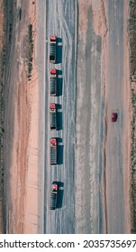 Convoy Of Trucks Rides On Asphalt Road With One Red Car On Another Side Of Road, Top View. Trucks Carry Building Materials For Construction Of The Road. Top Drone Shot.