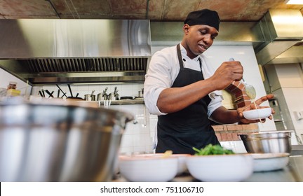 Cook Adding Sauce In Bowl At Commercial Kitchen. African Male Chef Cooking Food In Restaurant Kitchen.