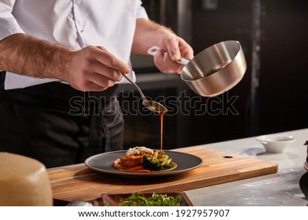 Cook in apron adding some sauce to dish. Cropped chef preparing food, meal, in kitchen, chef cooking, Chef decorating dish, closeup