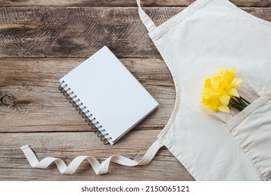 Cook Book With A Bouquet Of Fresh Daffodils On An Apron On A Wooden Background. Top View. Flat Lay