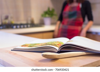 Cook Book Put On A Kitchen Table With A Wooden Spoon In The Foreground. In The Background You Can See A Young Woman With Apron Cooking In The Kitchen.