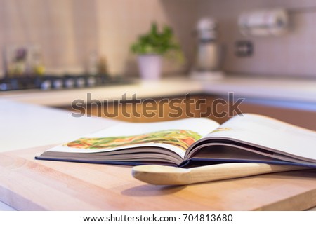 Cook book put on a kitchen table with a wooden spoon in the foreground. Kitchen is visible in the background.