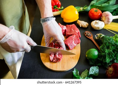 Cook Cutting Meat On A Board And Fresh Raw Vegetables On A Dark Table. Top View. Food Concept