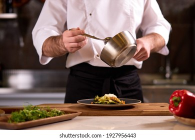 Cook dressing salad with sauce, pouring sauce into plate, finishing cooking of meal in restaurant kitchen, cropped man in apron at work - Shutterstock ID 1927956443