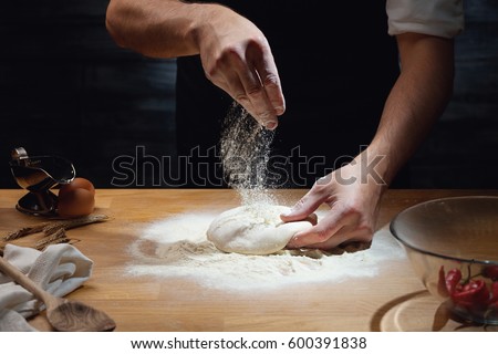 Cook hands kneading dough, sprinkling piece of dough with white wheat flour. Low key shot, close up on hands, some ingredients around on table.