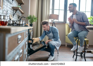 Cook at home. Young fair-haired man in an apron takes out a baking tray with pastries from the oven, a dark-haired man sitting next to him on a stool admiringly. - Powered by Shutterstock