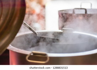 The Cook Is Opening The Lid Of The Large Soup Pot That Is Boiling With Steam.