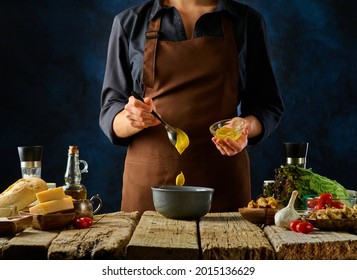 The cook prepares the sauce for the Caesar salad. He adds mustard to the bowl. The other ingredients for the salad lie side by side on a rough wooden table. Restaurant dish.