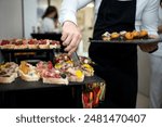 Cook puts snacks on table. Catering. Festive buffet. Table setting. Desserts and snacks on catering table. Waiter in black apron with tray. Bruschetta, desserts with fruit in baskets. Front view.