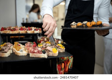 Cook puts snacks on table. Catering. Festive buffet. Table setting. Desserts and snacks on catering table. Waiter in black apron with tray. Bruschetta, desserts with fruit in baskets. Front view. - Powered by Shutterstock