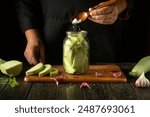 A cook spoons salt into a jar of chopped vegetable marrow before canning it with dill and garlic. Concept of pickling delicious zucchini on the kitchen table.
