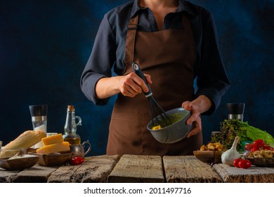 The Cook Whips The Sauce For Caesar Salad Dressing With A Hand Mixer. Cheese, Tomatoes, Eggs, Olive Oil And Other Ingredients For Caesar Salad Lie On A Wooden Table. Color Image. Blue Background.