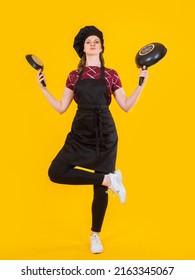 Cook Woman. Girl Cook Stands On One Leg. Confectioner With Frying Pans. Girl In Chef's Hat On Yellow. Female Student Learning To Cook. Concept Selling Frying Pans For Kitchen. Pastry Chef Career