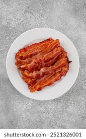 Cooked bacon for breakfast on a white plate on a gray concrete background. Top view, copy space