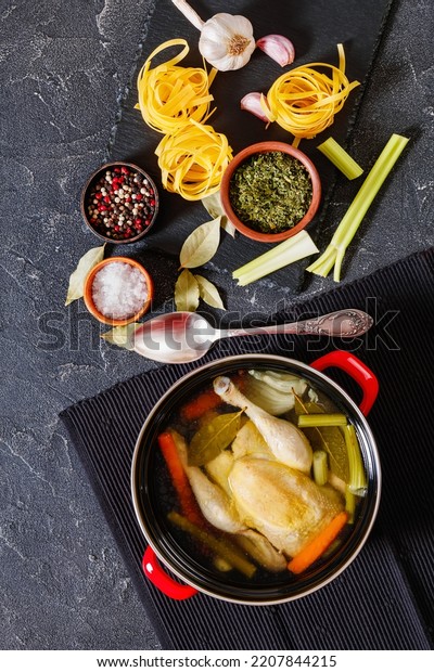 cooked chicken bouillon of whole\
chicken, celery, onion and carrot in red pot with ingredients and\
tagliatelle pasta on concrete table, vertical view from\
above