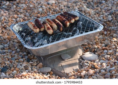 Cooked Chicken Meat Sausages Cooking On Instant Tin BBQ Grill Browned And Burnt From Hot Charcoal Fire At Torcross Surrounded By Natural Beach Pebbles And Stones On Top Of Slate On Summer Day