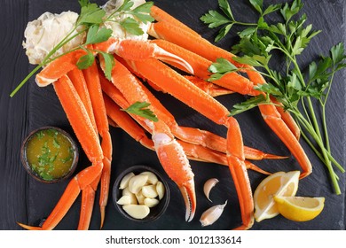 Cooked Crab Legs With Melted Butter, Garlic Cloves, Lemon Slices And Fresh Parsley On Black Stone Tray, On Wooden Table, View From Above, Close-up