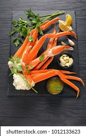 Cooked Crab Legs With Melted Butter, Garlic Cloves, Lemon Slices And Fresh Parsley On Black Stone Tray, On Wooden Table, Vertical View From Above, Close-up