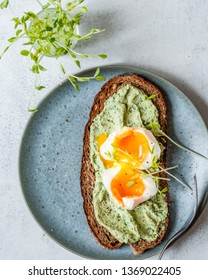A cooked egg on toast of a grain bread with a sauce with herbs and fresh microgreen pea and radish sprouts. View from above. Healthy breakfast on a gray-blue ceramic plate on a light stone background.