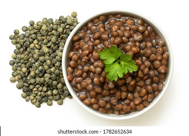 Cooked French Green Puy Lentils With Garnish In White Ceramic Bowl Next To Dry Lentils Isolated On White. Top View.