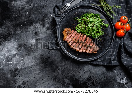 Cooked grilled strip loin steak, marbled beef meat with arugula. Black background. Top view. Copy space.