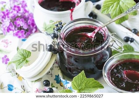 Cooked homemade black currant jam in jar on white wooden table and vivid tablecloth background, fresh black currant jam close up, food and healthy eating concept