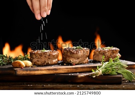 Cooked juicy steak meat beef with hand sprinking seasoning on top on wooden chopping board with flames in the background.
