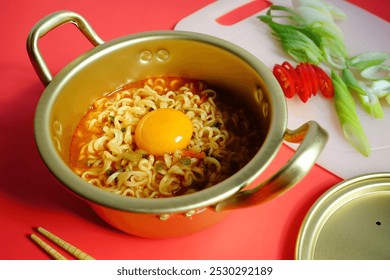 Cooked ramyeon or Korean instant noodles soup with egg in double handle Korean yellow aluminum pot on red background with chopsticks and white plastic cutting board with sliced red chili, scallion. - Powered by Shutterstock