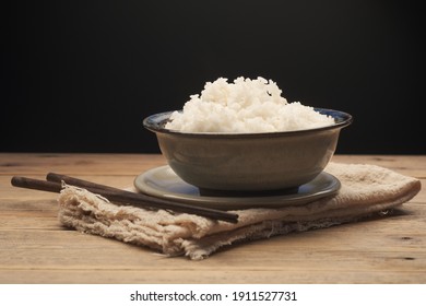 Cooked Thai jasmine rice in a ceramic bowl with chopsticks placed on an old wooden table against a black background.