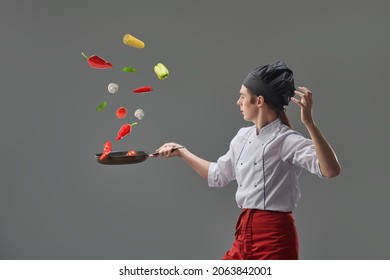 Cooking art. A professional young chef in neat uniform demonstrates his cooking art tossing vegetables in a frying pan. Sideview portrait. Gray studio background.  - Powered by Shutterstock