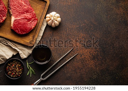 Cooking background for menu or template with raw meat steaks on wooden cutting board, spices and olive oil on rustic stone background with space for text 