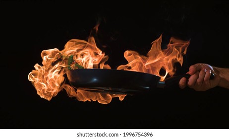 Cooking By A Chef, Processing Food With A Flame In A Frying Pan On A Black Background,  The Concept Of Restaurant Service, Impressive Cooking