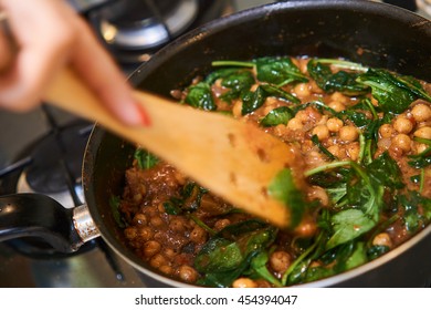 Cooking Chickpeas With Spinach In A Pan, On A Stove, Using A Wooden Spoon